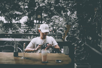 Young man holding camera while sitting on table