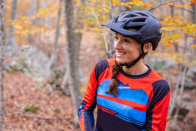 Delighted woman in helmet and sportswear smiling and looking away while cycling in autumn forest