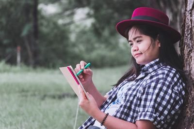 Side view of young woman writing on book while sitting at park