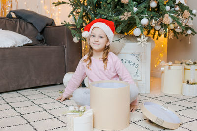 Happy girl in pajamas sits with gift in santa claus hat in room decorated