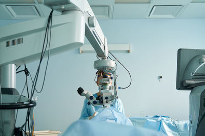 Attentive female doctor in surgical uniform and sterile mask looking through microscope while operating eye of unrecognizable patient in hospital