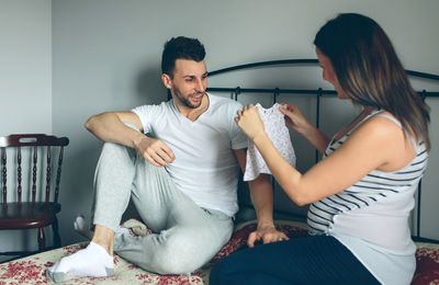 Couple looking baby clothing while sitting on bed at home