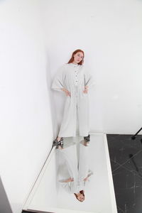 Full length fashion catalog style portrait of woman in jumpsuit standing on mirror over white wall