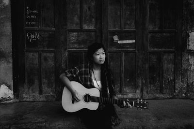 Young woman playing guitar while sitting against old wall