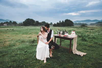 Full length of wedding couple sitting on chair at farm against cloudy sky