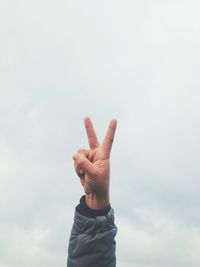 Cropped image of hand making peace sign against cloudy sky