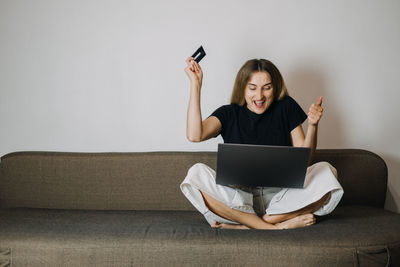 Full length of cheerful woman holding credit card while using laptop