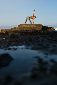 Side view of slim female in sportswear doing revolved triangle asana while practicing yoga on rocky seashore