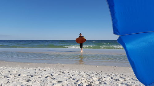 Full length of young man holding surfboard while walking at beach against clear sky