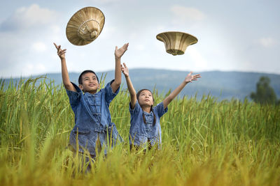 Siblings throwing hats while standing on field against sky