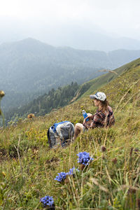 Young woman sitting on meadow in mountain valley next to backpack drinking water  on hike landscape 