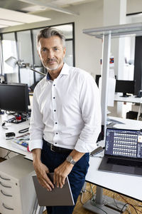 Confident businessman with digital tablet staring while standing by desk at office