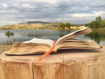 Open book on wood by lake against cloudy sky