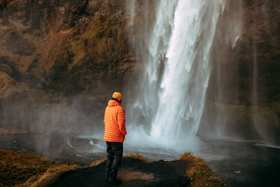 Back view of young tourist in winter wear looking at cascade and mountain river between stone hill