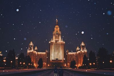 Rear view of young couple standing near illuminated historic buildings during snowfall at night