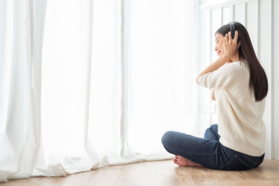 Side view of woman sitting on floor against window