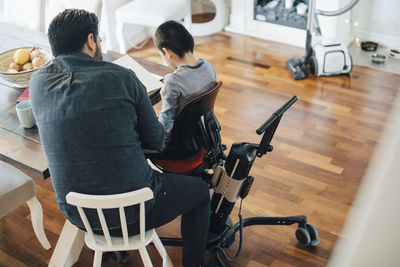 High angle view of father teaching autistic son sitting on wheelchair in living room