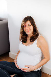 Thoughtful pregnant woman sitting against white wall at home