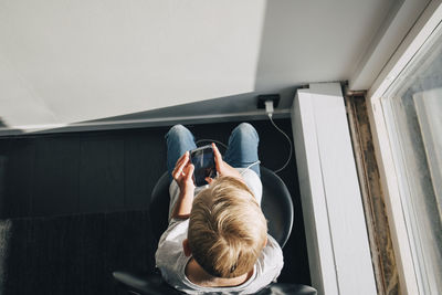 High angle view of boy using smart phone sitting on chair by window at home