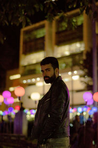 Portrait of young man standing by illuminated buildings in city night