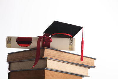 Close-up of diploma and mortarboard on stacked books against white background