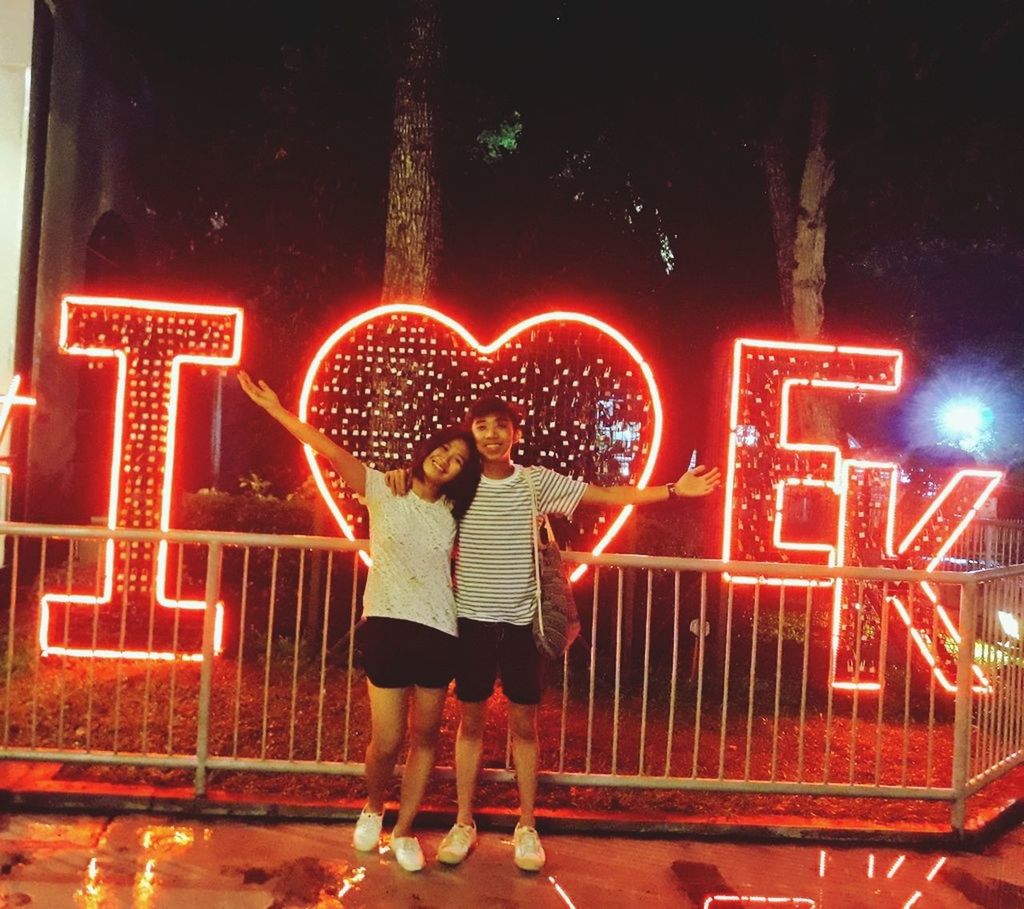 illuminated, love, full length, western script, text, heart shape, real people, glowing, two people, front view, communication, night, red, casual clothing, standing, togetherness, outdoors, childhood, lifestyles, alphabet, bonding, neon, people