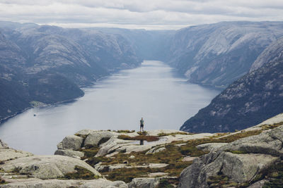 Distant view of hiker standing on rock by river and mountains in foggy weather