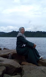 Portrait of smiling young woman sitting on rock by lake against sky