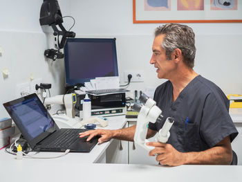 Side view of mature man with microscope browsing laptop while working in modern medical laboratory in hospital