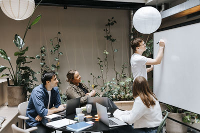 Young male entrepreneur writing on whiteboard in meeting with colleagues at office
