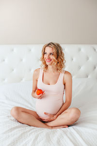 Pregnant smiling woman holding fruit while sitting on bed at home