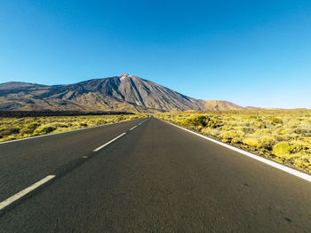 Road leading towards mountains against clear blue sky