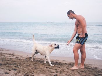 Side view of man playing with dog at shore against sky