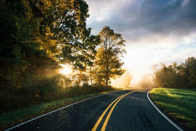 Trees growing by empty road against sky during sunrise