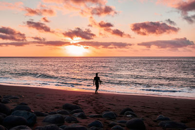 Silhouette of distant anonymous tourist scrolling on sandy coast near waving sea at sundown time during summer vacation in tropical resort