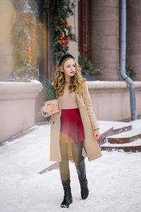 A young hipster woman walking through the city's snow-covered streets holds a potted christmas tree