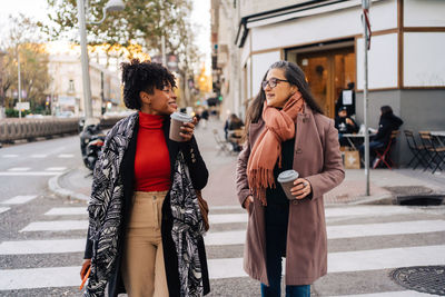 Content diverse female friends in warm clothes with coffee to go looking at each other while crossing city street