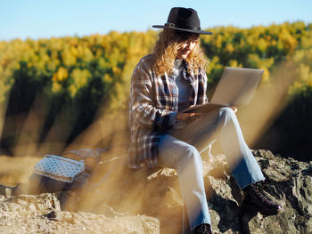 Young curly woman freelancer in felt hat and plaid t-shirt using laptop at mountain in autumn