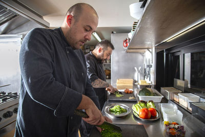 Side view of professional male cooks in uniforms cutting greens and vegetable while preparing food at counter in light kitchen of modern restaurant