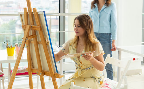 Young woman painting while woman standing in background