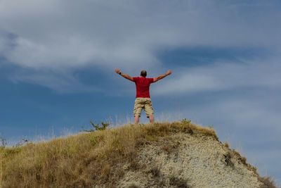 Low angle view of man standing with arms outstretched on hill against sky