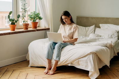 Young female brunette sitting in the room on the bed by the window working on a portable laptop