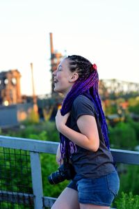 Side view of smiling girl carrying camera while leaning on railing