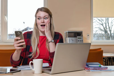 Shocked businesswoman using mobile phone while sitting by laptop in office