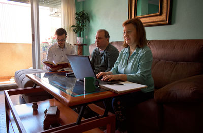 Father watch tv, son reads a book and mom works on her laptop