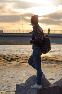 Side view of woman standing by railing against sky during sunset