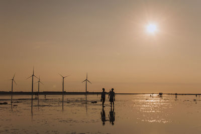 Silhouette people standing by windmills at wet beach during sunset