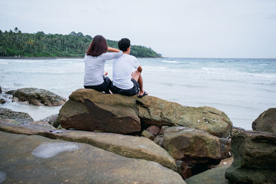 Rear view of couple sitting on rock against sea