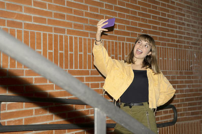 Smiling young woman doing selfie while standing against wall