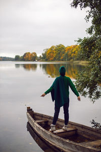 Woman in a green sweater stands with her back in an old wooden boat on a lake in autumn